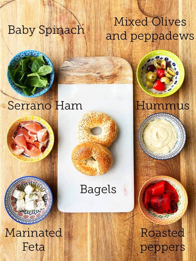 Two toasted bagels rest on a marble board, surrounded by colourful bowls of various leftover antipasti on a wooden board.