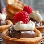 Golden brown Nutella tartlets with puff pastry, topped with whipped cream, a raspberry, and crushed hazelnuts, displayed on a wire rack.