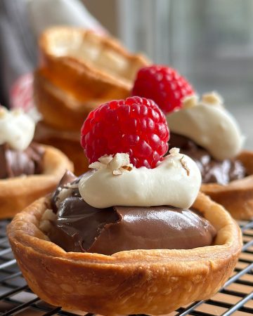 Golden brown Nutella tartlets with puff pastry, topped with whipped cream, a raspberry, and crushed hazelnuts, displayed on a wire rack.