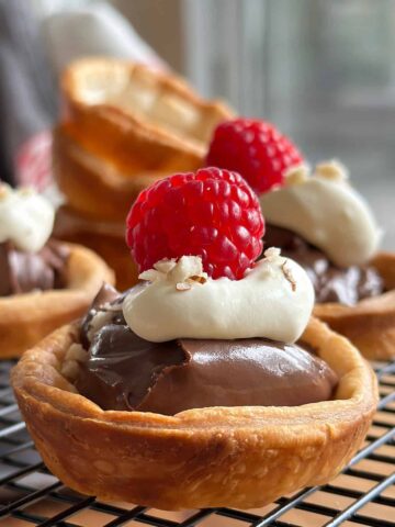 Golden brown Nutella tartlets with puff pastry, topped with whipped cream, a raspberry, and crushed hazelnuts, displayed on a wire rack.