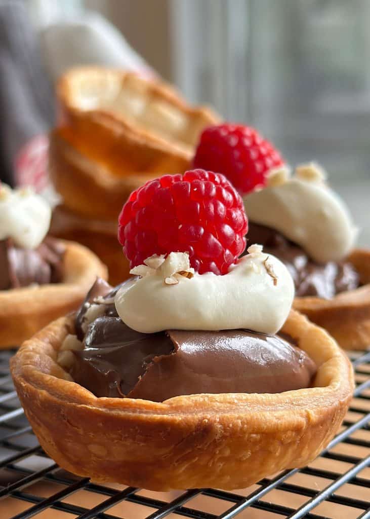 Golden brown Nutella tartlets with puff pastry, topped with whipped cream, a raspberry, and crushed hazelnuts, displayed on a wire rack.