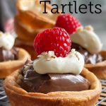 Golden brown Nutella tartlets with puff pastry, topped with whipped cream, a raspberry, and crushed hazelnuts, displayed on a wire rack.