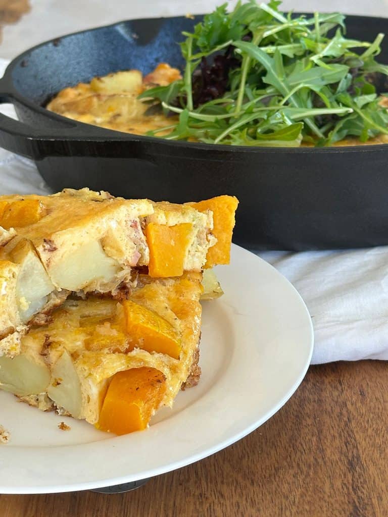 Two slices of pumpkin frittata on a white plate, with a skillet containing the baked frittata topped with leaves in the background.