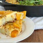 Two slices of pumpkin frittata on a white plate, with a skillet containing the baked frittata topped with leaves in the background.