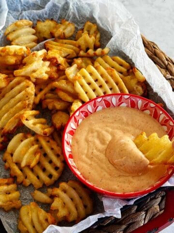 A rustic plate of waffle fries with a bowl of honey BBQ aioli on the side and a hand dipping a fry into the sauce.