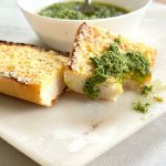 Basil pesto or pesto al basilico in a white bowl with two slices of garlic bread on a marble board, one slice topped with a dollop of pesto and a bite taken out of it.