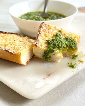 Basil pesto or pesto al basilico in a white bowl with two slices of garlic bread on a marble board, one slice topped with a dollop of pesto and a bite taken out of it.