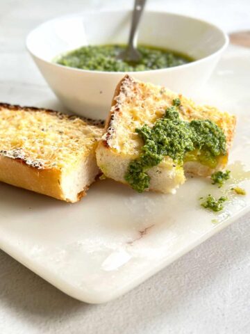 Basil pesto or pesto al basilico in a white bowl with two slices of garlic bread on a marble board, one slice topped with a dollop of pesto and a bite taken out of it.