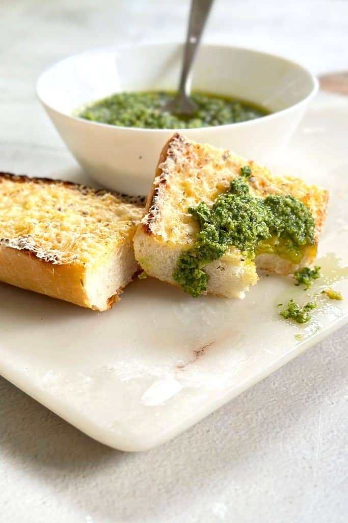 Basil pesto or pesto al basilico in a white bowl with two slices of garlic bread on a marble board, one slice topped with a dollop of pesto and a bite taken out of it.