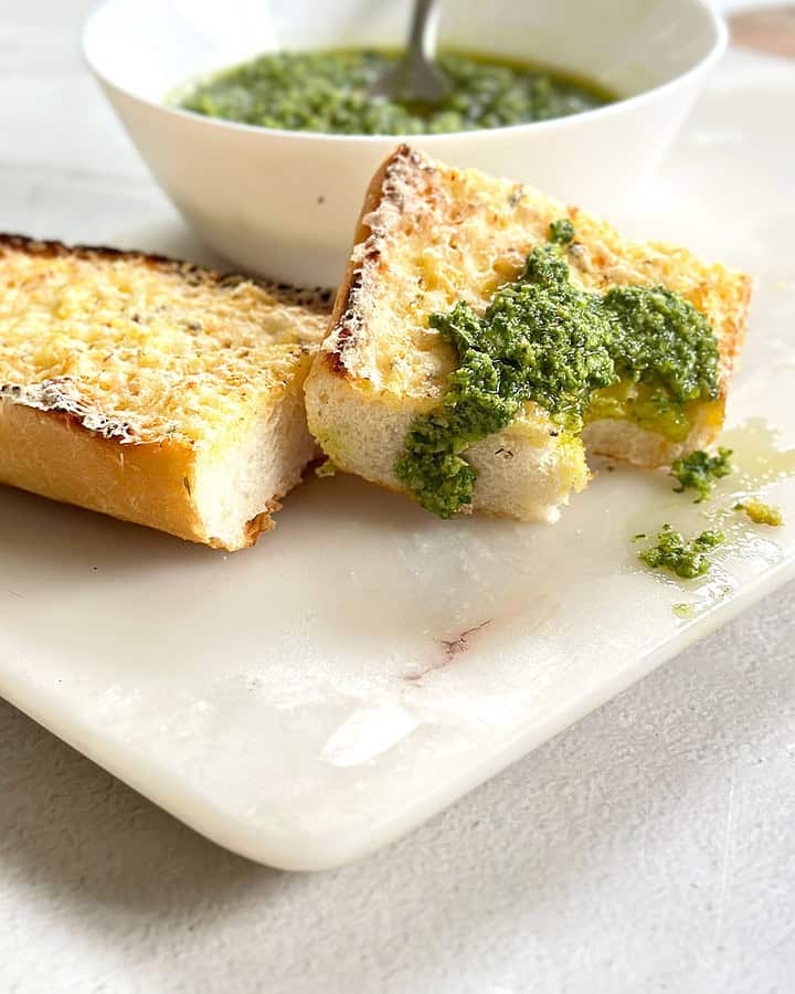 Basil pesto or pesto al basilico in a white bowl with two slices of garlic bread on a marble board, one slice topped with a dollop of pesto and a bite taken out of it.