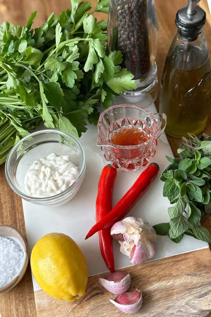 All the ingredients for Chimichurri mayonnaise are arranged on a wooden board, including coriander, oregano, parsley, oil, chili, lemon, red wine vinegar, garlic, mayonnaise, and salt and pepper.