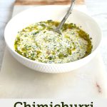 A white bowl filled with Chimichurri mayo, showing specks of green fresh herbs and red chilli mixed into a creamy white sauce, placed on a marble serving board.