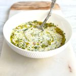 A white bowl filled with chimichurri mayo, showing specks of green fresh herbs and red chili mixed into a creamy white sauce, placed on a marble serving board.