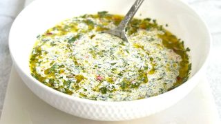 A white bowl filled with chimichurri mayo, showing specks of green fresh herbs and red chili mixed into a creamy white sauce, placed on a marble serving board.