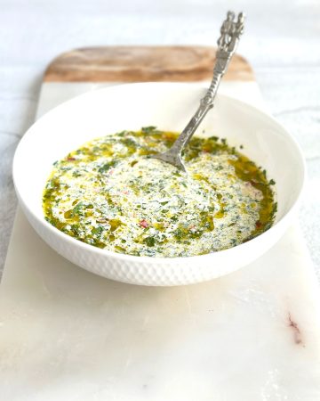 A white bowl filled with chimichurri mayo, showing specks of green fresh herbs and red chili mixed into a creamy white sauce, placed on a marble serving board.