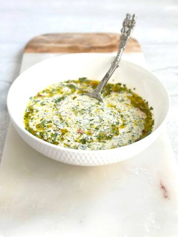 A white bowl filled with chimichurri mayo, showing specks of green fresh herbs and red chili mixed into a creamy white sauce, placed on a marble serving board.