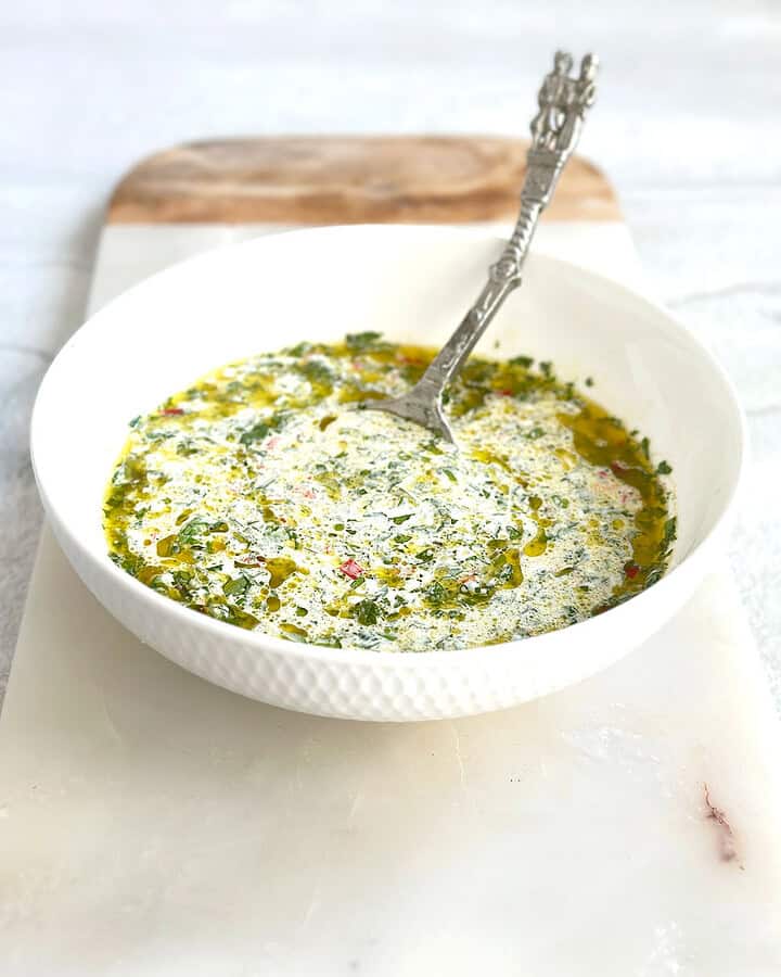 A white bowl filled with chimichurri mayo, showing specks of green fresh herbs and red chili mixed into a creamy white sauce, placed on a marble serving board.