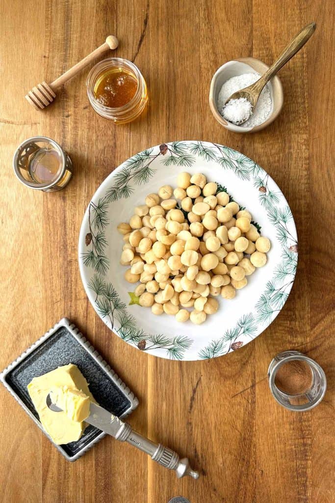 Ingredients for honey roasted macadamia nuts arranged on a wooden board, including butter, macadamia nuts, cinnamon, vanilla, sea salt, and honey.