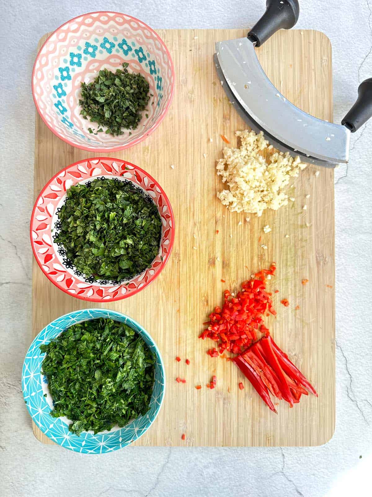 Three small bowls of finely chopped parsley, coriander, and oregano, with finely chopped red chilli and garlic on the side, arranged on a wooden cutting board with a herb chopping tool in the background.