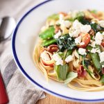 A white bowl with blue rim filled with pasta with pancetta and peas, topped with torn basil leaves, spinach, cherry tomatoes, and crumbled feta.