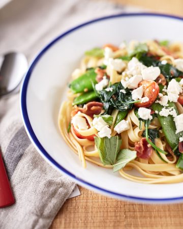 A white bowl with blue rim filled with pasta with pancetta and peas, topped with torn basil leaves, spinach, cherry tomatoes, and crumbled feta.