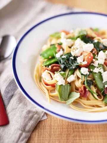 A white bowl with blue rim filled with pasta with pancetta and peas, topped with torn basil leaves, spinach, cherry tomatoes, and crumbled feta.