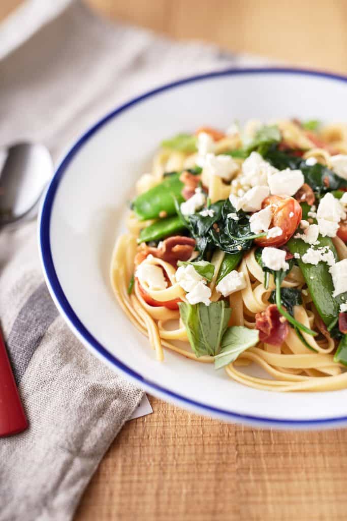 A white bowl with blue rim filled with pasta with pancetta and peas, topped with torn basil leaves, spinach, cherry tomatoes, and crumbled feta.