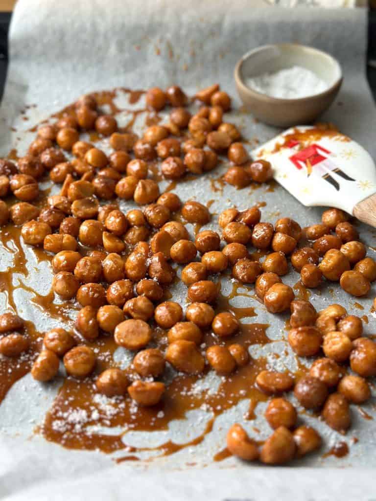 Deep caramel-coloured honey roasted macadamia nuts evenly spread on a baking sheet, lightly dusted with sea salt.