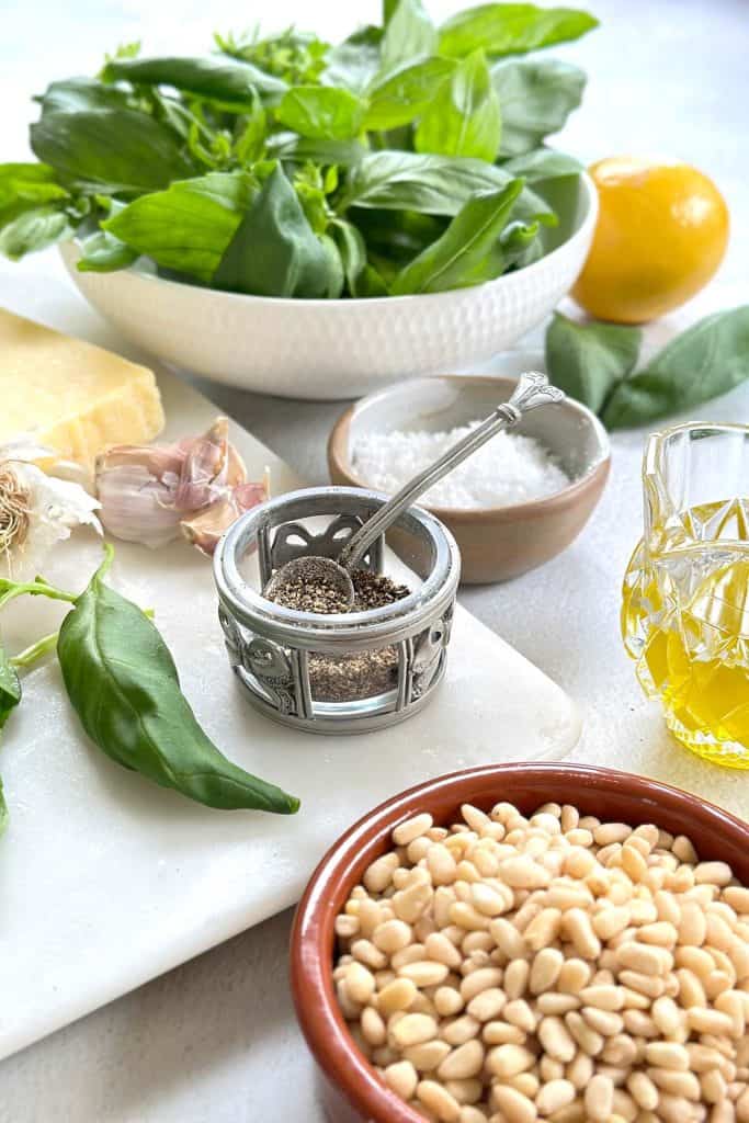 All the ingredients for traditional pesto alla Genovese are arranged on a white background, including a white bowl with fresh basil, a piece of Parmesan cheese, a bowl of pine nuts, a lemon, olive oil, garlic, salt, and pepper.