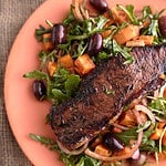 Juicy steak served on a bed of arugula steak salad with roasted sweet potato cubes, olives, and red onion, all plated on a vibrant orange dish.