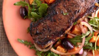 Juicy steak served on a bed of arugula steak salad with roasted sweet potato cubes, olives, and red onion, all plated on a vibrant orange dish.