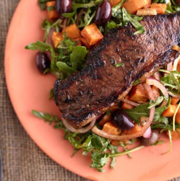 Juicy steak served on a bed of arugula steak salad with roasted sweet potato cubes, olives, and red onion, all plated on a vibrant orange dish.