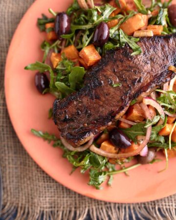 Juicy steak served on a bed of arugula steak salad with roasted sweet potato cubes, olives, and red onion, all plated on a vibrant orange dish.