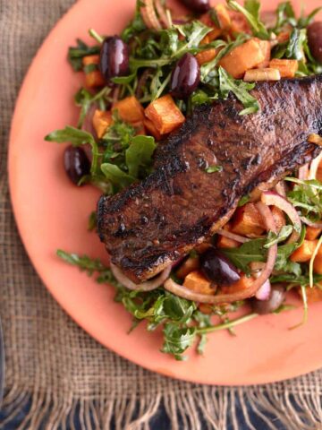 Juicy steak served on a bed of arugula steak salad with roasted sweet potato cubes, olives, and red onion, all plated on a vibrant orange dish.