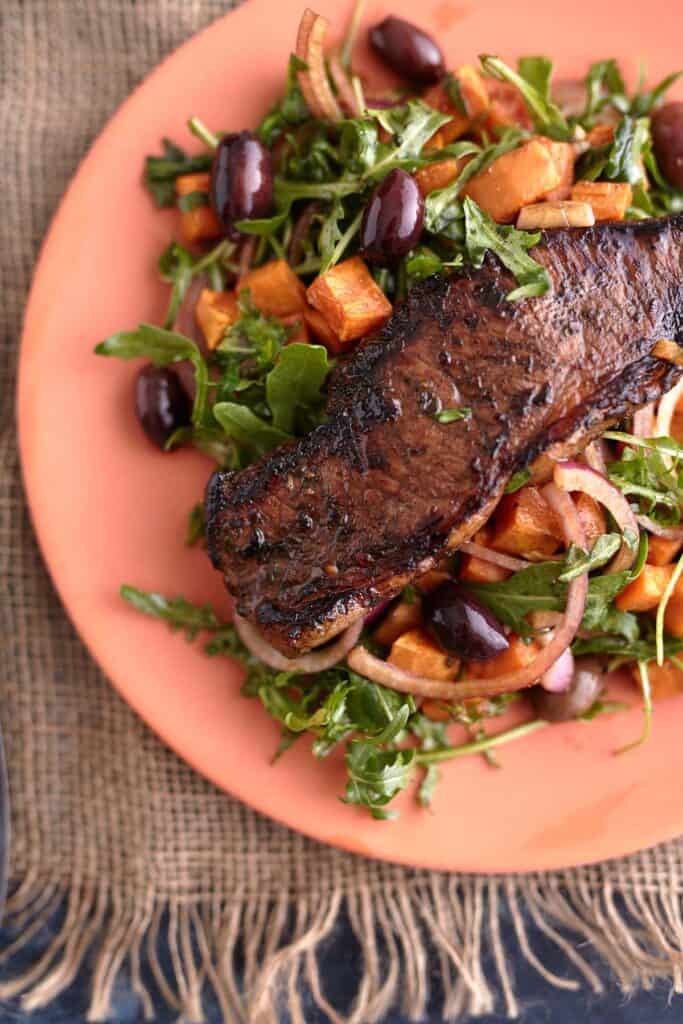 Juicy steak served on a bed of arugula steak salad with roasted sweet potato cubes, olives, and red onion, all plated on a vibrant orange dish.