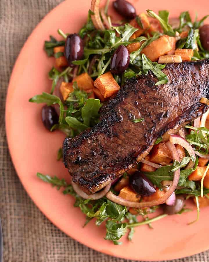 Juicy steak served on a bed of arugula steak salad with roasted sweet potato cubes, olives, and red onion, all plated on a vibrant orange dish.