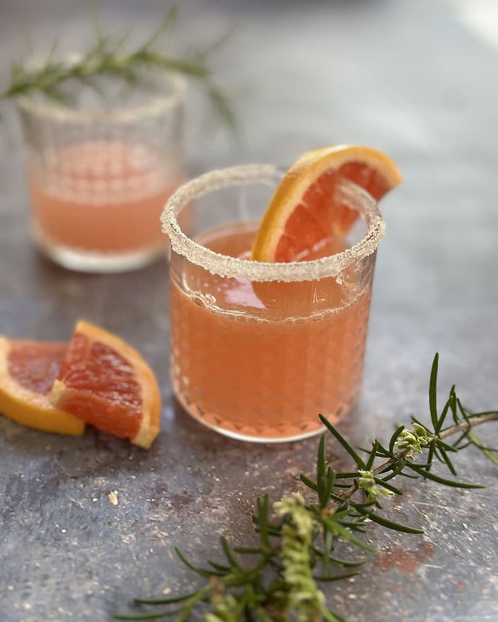 Two sugar-rimmed glasses filled with pink grapefruit gin cocktails, garnished with fresh rosemary sprigs and slices of grapefruit.