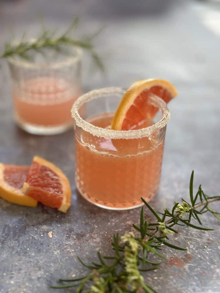 Two sugar-rimmed glasses filled with pink grapefruit gin cocktails, garnished with fresh rosemary sprigs and slices of grapefruit.