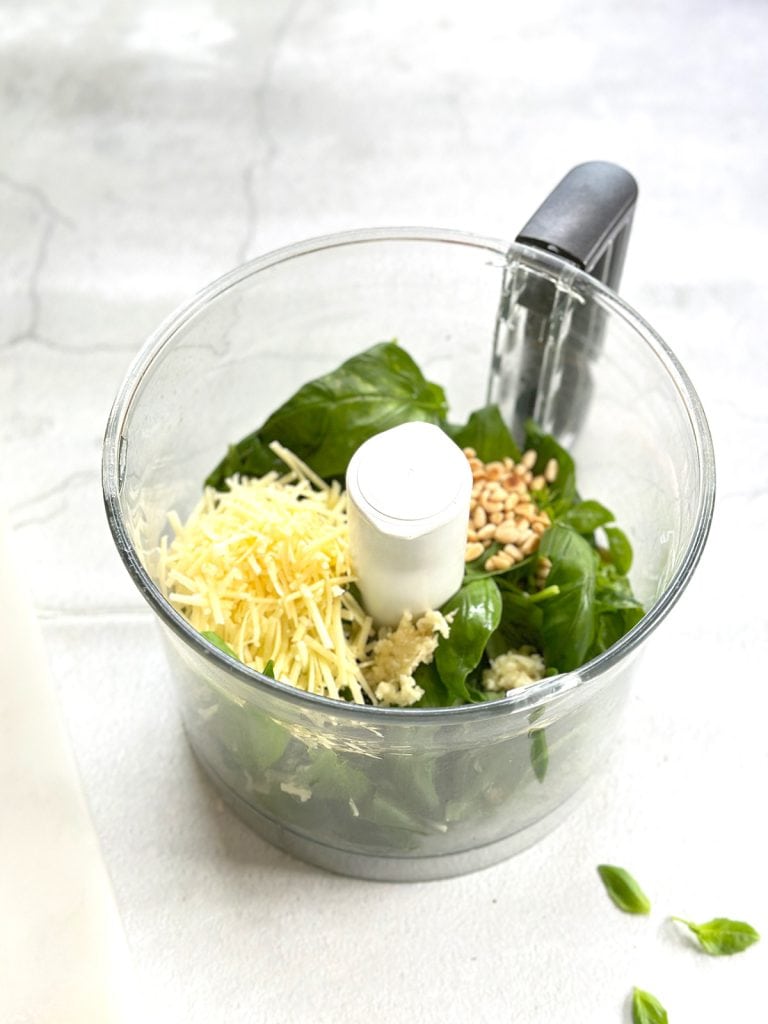 All the ingredients for pesto alla Genovese—grated parmesan, toasted pine nuts, garlic, and basil—are in a food processor, ready to be pulsed.