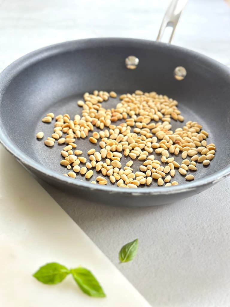 Toasted pine nuts sit in a black frying pan with fresh basil leaves scattered on a white marble surface.