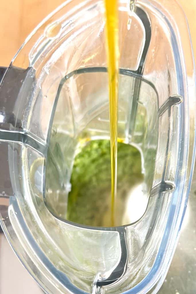 A slow, steady stream of olive oil is added to a food processor while it is running.