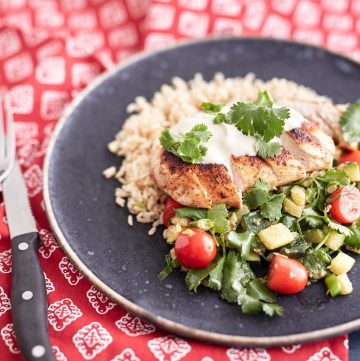 Sliced crispy chicken served over a bed of brown rice, accompanied by a zucchini and cherry tomato salad, topped with a dollop of sour cream and garnished with fresh coriander leaves.