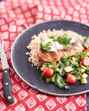 Sliced crispy chicken served over a bed of brown rice, accompanied by a zucchini and cherry tomato salad, topped with a dollop of sour cream and garnished with fresh coriander leaves.