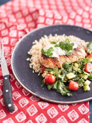 Sliced crispy chicken served over a bed of brown rice, accompanied by a zucchini and cherry tomato salad, topped with a dollop of sour cream and garnished with fresh coriander leaves.