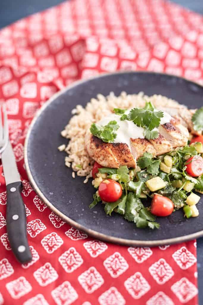 Sliced crispy chicken served over a bed of brown rice, accompanied by a zucchini and cherry tomato salad, topped with a dollop of sour cream and garnished with fresh coriander leaves.