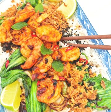 A plate of tiger prawns and bok choy on a bed of jasmine rice, topped with crispy chili onion and black sesame seeds, served with a lime wedge on the side.