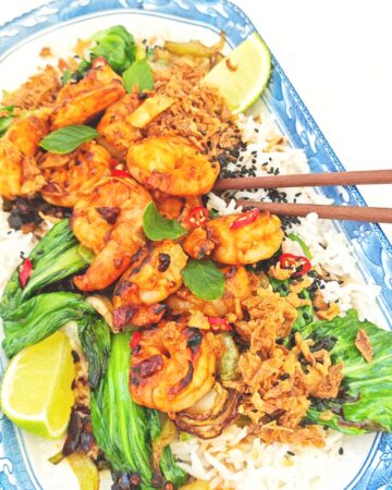 A plate of tiger prawns and bok choy on a bed of jasmine rice, topped with crispy chili onion and black sesame seeds, served with a lime wedge on the side.