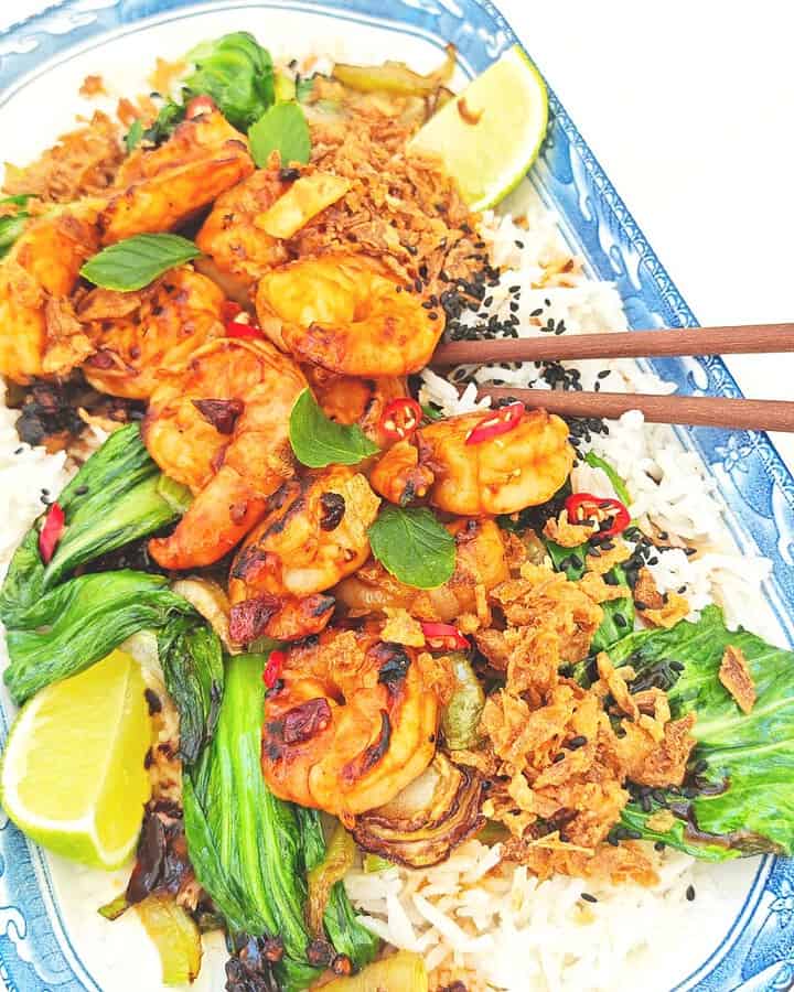 A plate of tiger prawns and bok choy on a bed of jasmine rice, topped with crispy chili onion and black sesame seeds, served with a lime wedge on the side.