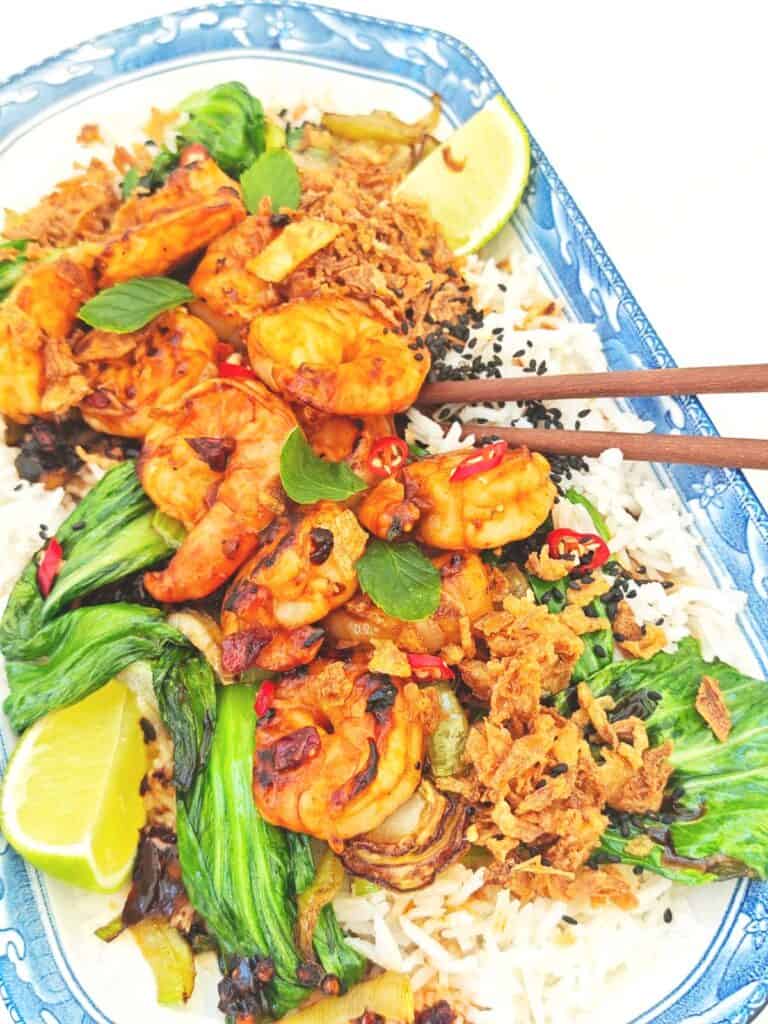 A plate of tiger prawns and bok choy on a bed of jasmine rice, topped with crispy chili onion and black sesame seeds, served with a lime wedge on the side.