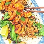 A plate of tiger prawns and bok choy on a bed of jasmine rice, topped with crispy chili onion and black sesame seeds, served with a lime wedge on the side.